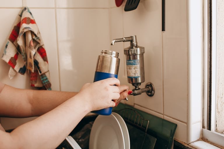 Close-up of hands filling a reusable water bottle at an indoor kitchen sink.