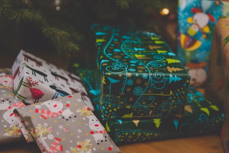Christmas gifts in natural materials under a lit tree: a wooden toy train and blocks, folded wool blankets, a ceramic pot, a lit candle and kraft paper parcels tied with twine