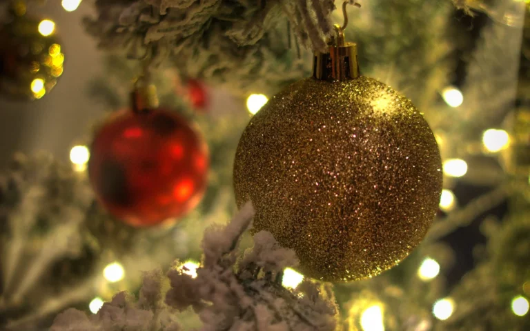 Close-up of a gold glitter-covered Christmas bauble hanging on a flocked tree branch, with warm fairy lights glowing behind it