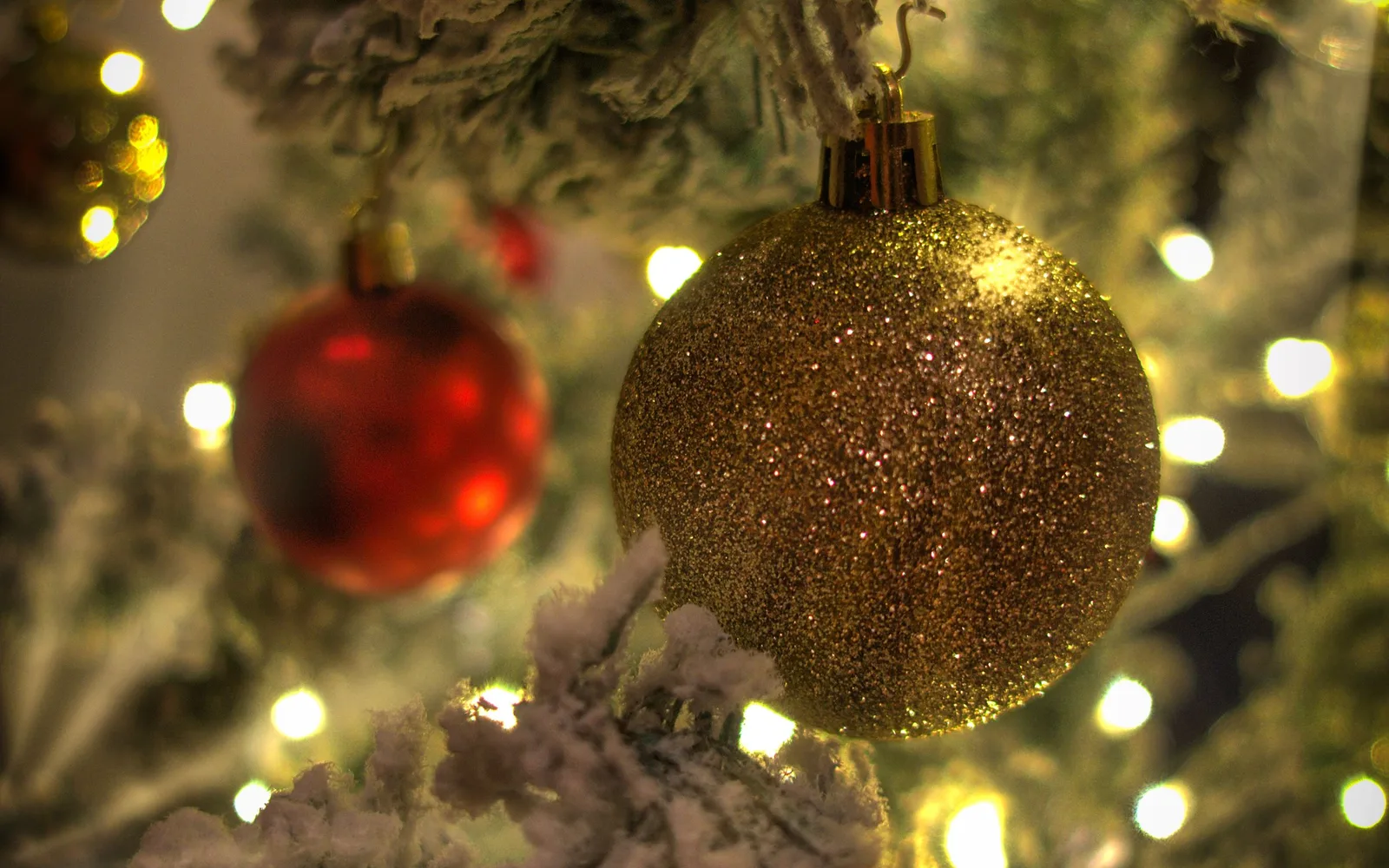 Close-up of a gold glitter-covered Christmas bauble hanging on a flocked tree branch, with warm fairy lights glowing behind it