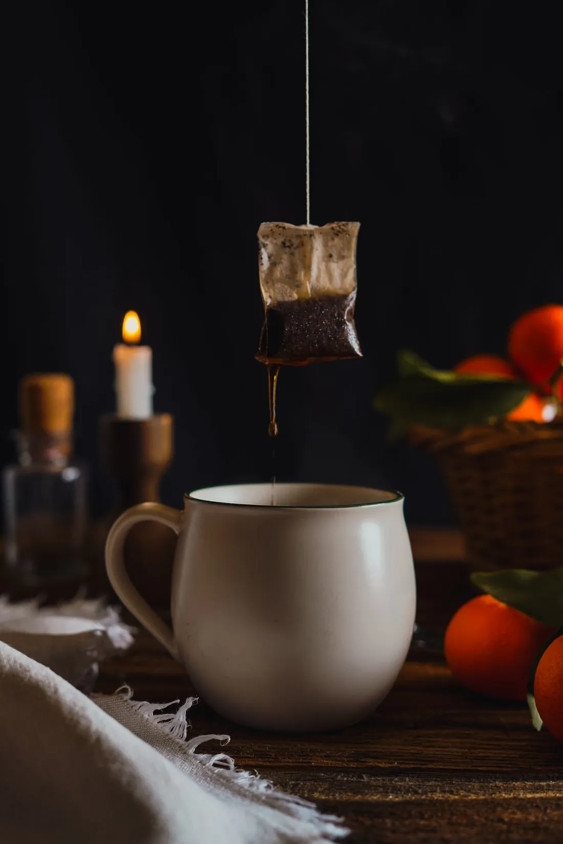 A tea bag dripping into a ceramic mug beside a lit candle and fresh oranges on a wooden table