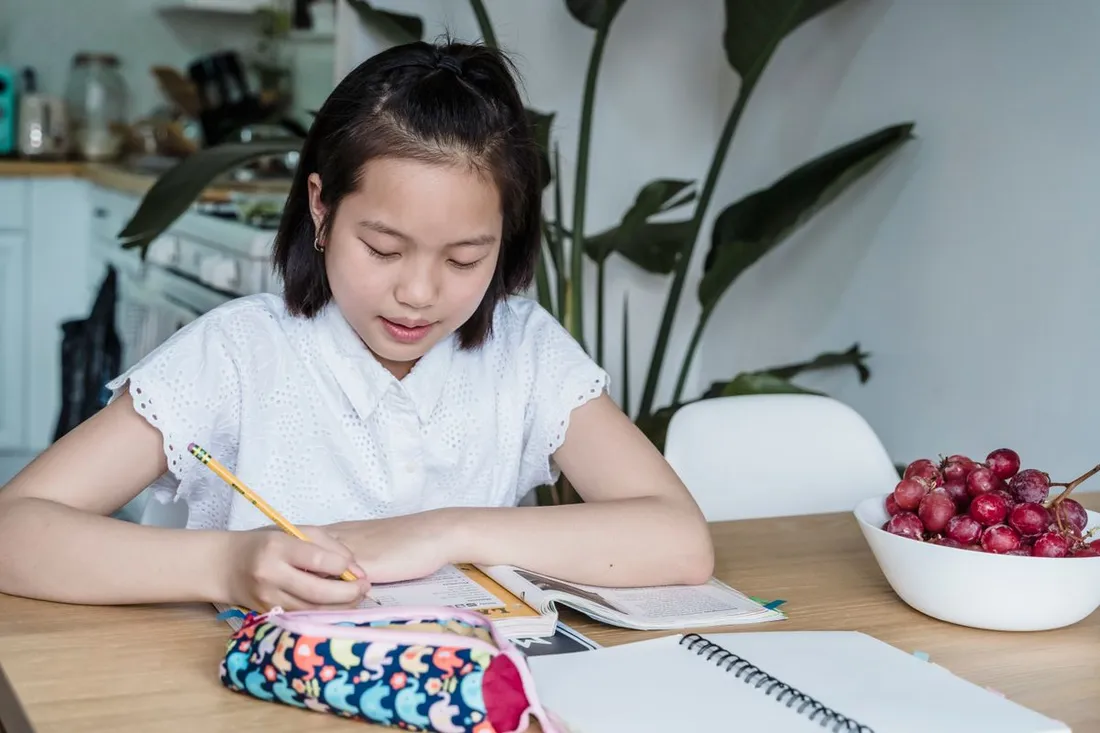 A teenager writing in a notebook at a table with a pencil case and a spiral notebook