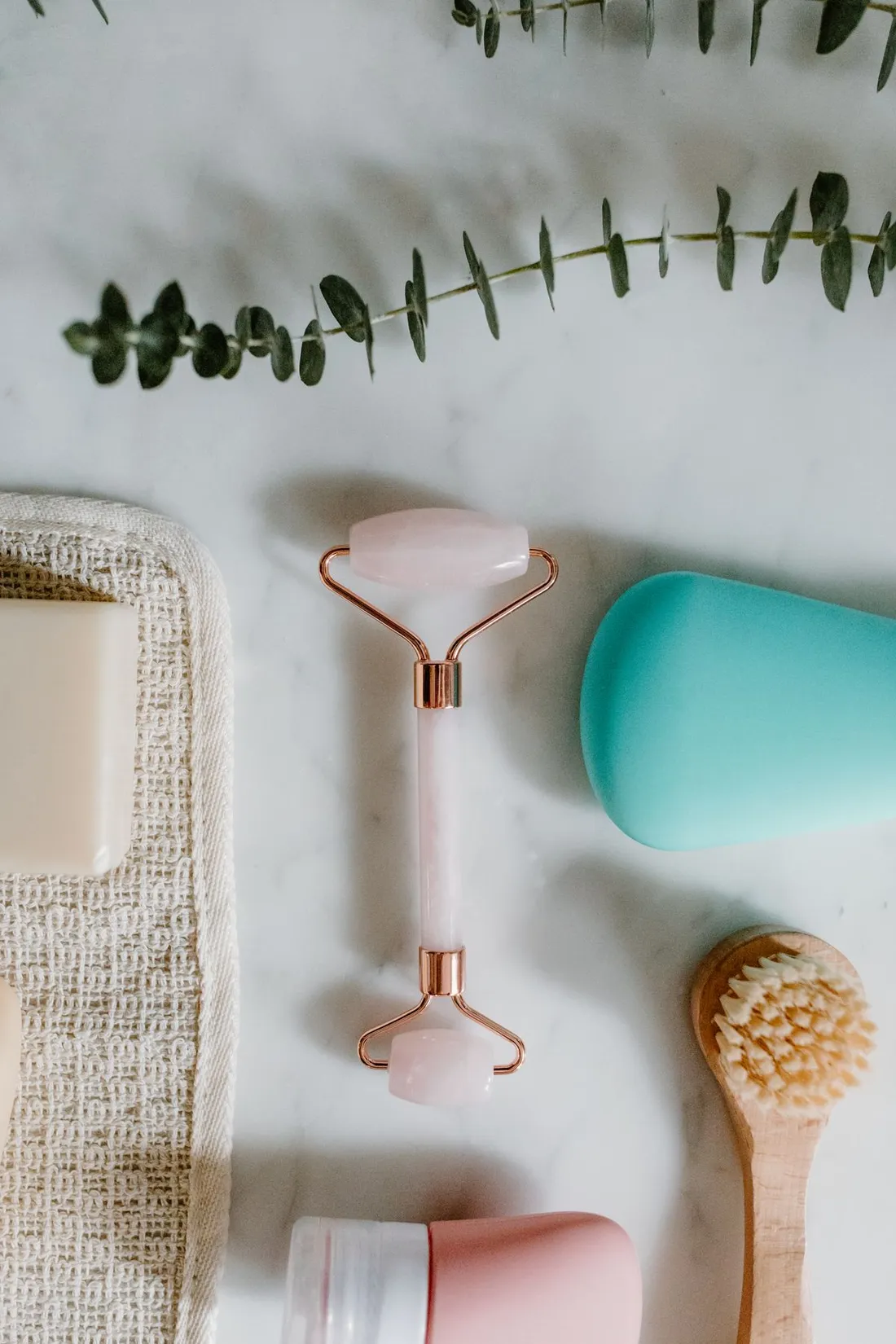 Natural self-care flat lay on marble: a stone face roller, a bar of soap, a wooden body brush and eucalyptus